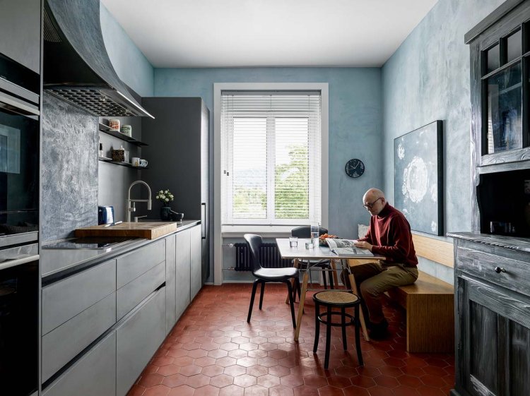 Ein Foto einer Küche. Ind er Küche befindet sich ein kleiner Tisch, der von unterschiedlichen Hockern umgeben ist. Der Besitzer der Wohnung sitzt auf der Sitzbank und schaut eine Zeitung an.