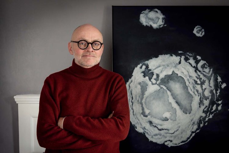 Ein Portrait-Foto des Künstlers Jean-Pierre Lanz. Er trägt eine runde Brille, einen roten Rollkragenpullover und steht vor einem weiteren Kunstwerk.