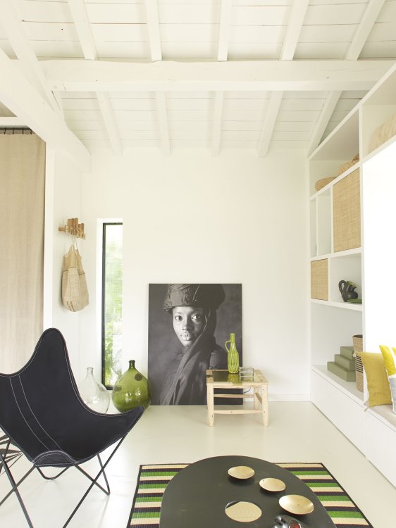 Weiss gestrichenes Wohnzimmer mit schwarzem Campingstuhl, einem ovalen Couchtisch auf einem grün-gelb-gestreiften Teppich und einem Schwarz-Weiss-Portrait einer Frau im Hintergrund.