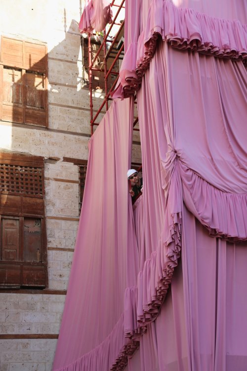 Der Künstler und Designer Andrés Reisinger guckt mit einem Baustellenhelm auf dem Kopf, zwischen den Stoffbahnen seiner Installation «Take Over Jeddah» hervor.